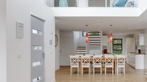 Un interior moderno con una zona de comedor que incluye una mesa de madera y sillas blancas, una escalera de peldaños de madera, y una cocina con armarios blancos.