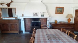 Uma sala de jantar com toalha xadrez, cadeiras de madeira, lareira e decoração rústica, incluindo um espelho e um quadro emoldurado.