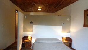 Quarto com teto de madeira, cama com colcha branca, duas mesas de cabeceira com abajures e um pequeno espelho na parede.