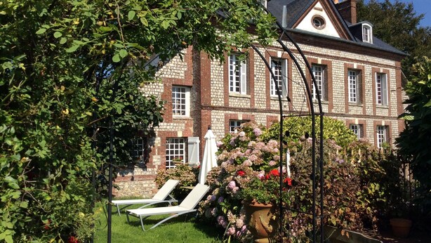 Un grande edificio in pietra con un giardino, due lettini bianchi e un vaso di fiori capiente.