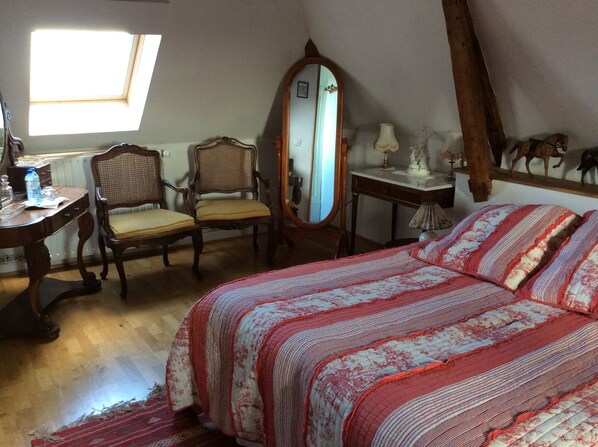 A bedroom with a large bed, a wooden chair, a desk with a mirror, and a small table with decorative items.