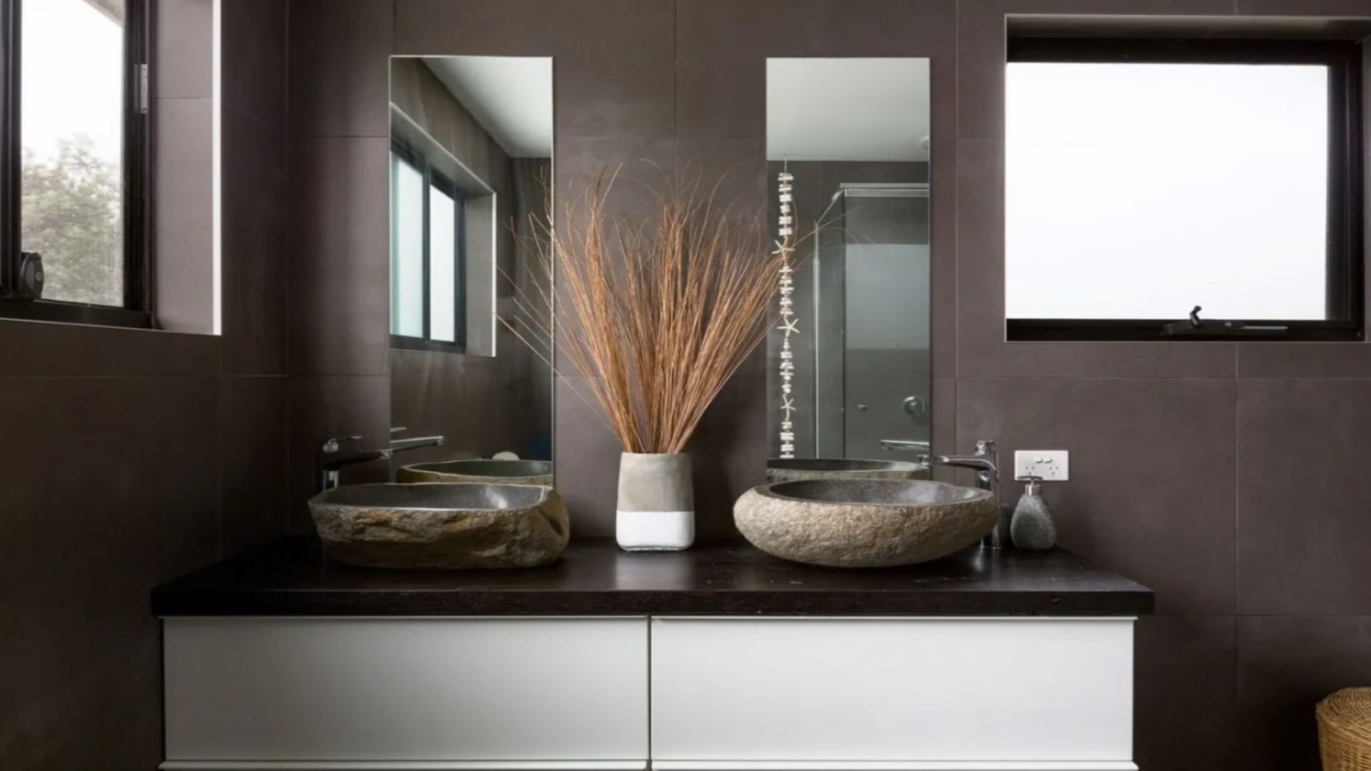A modern bathroom with two stone sinks, a tall mirror, and a decorative vase with reeds.