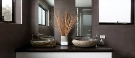 A modern bathroom with two stone sinks, a tall mirror, and a decorative vase with reeds.
