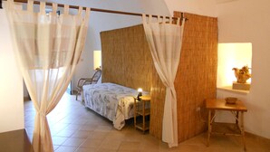 Un dormitorio con una cama, una silla, una mesa y una lámpara. Se ve una ventana con cortinas y una pared con un patrón texturizado.