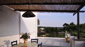 Un área de comedor al aire libre techada, con una mesa y sillas, una lámpara colgante y vistas a un jardín y edificios a lo lejos.