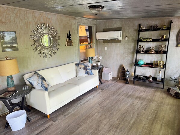 A living room with a white sofa, a sunburst mirror, a wall-mounted air conditioner, and a shelf with decorative items.