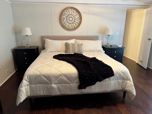 A neatly made bed with a white duvet and a black throw blanket, flanked by two bedside tables with lamps, a wall-mounted decorative piece, and a door leading to another room.