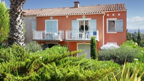 Una casa de dos pisos con tejado de tejas rojas, contraventanas blancas y un balcón con barandillas blancas.