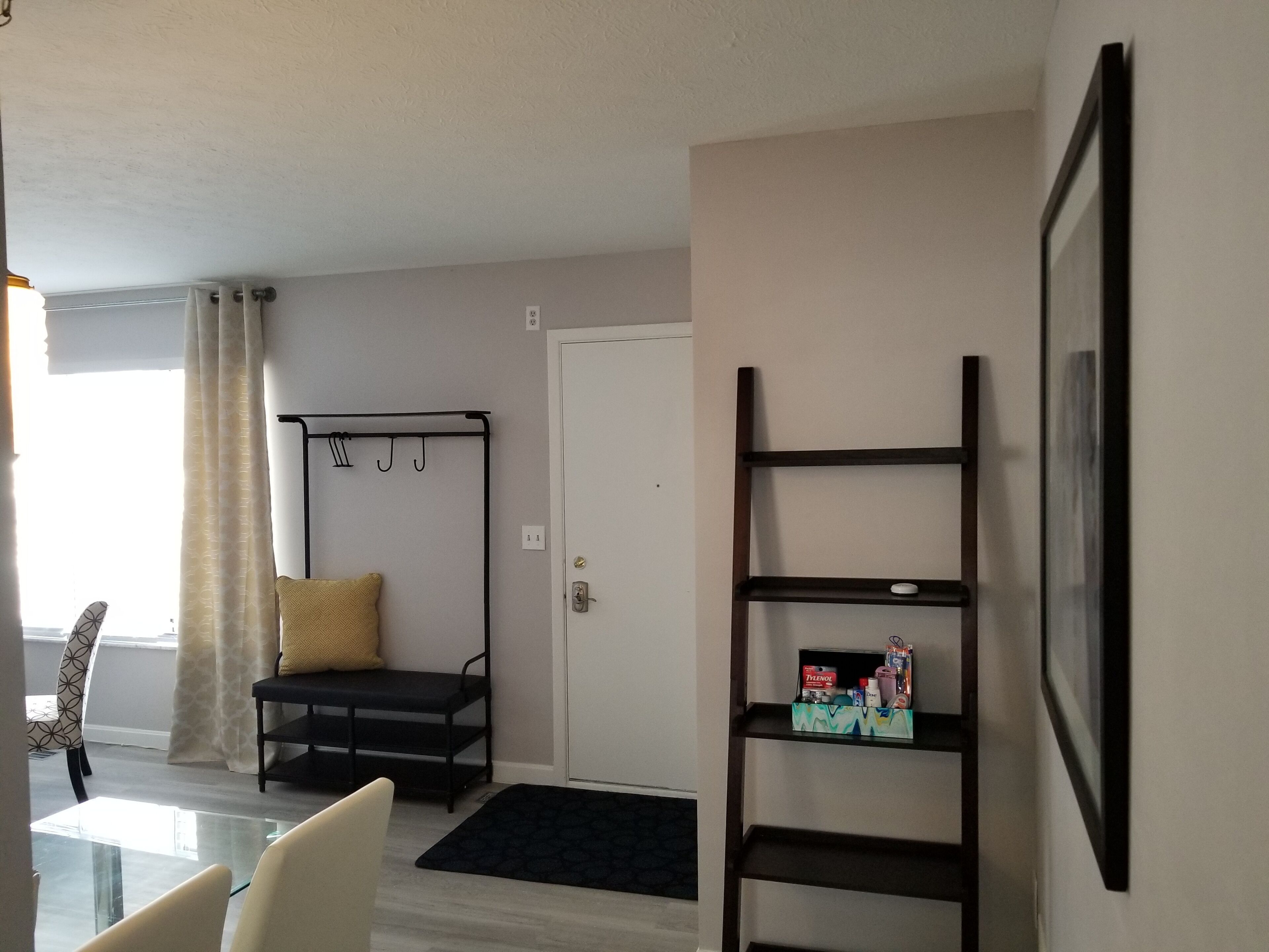 A modern living room with a black metal bench, a glass coffee table, and a black rug. There is a white door and a wall-mounted shelf with items on it.