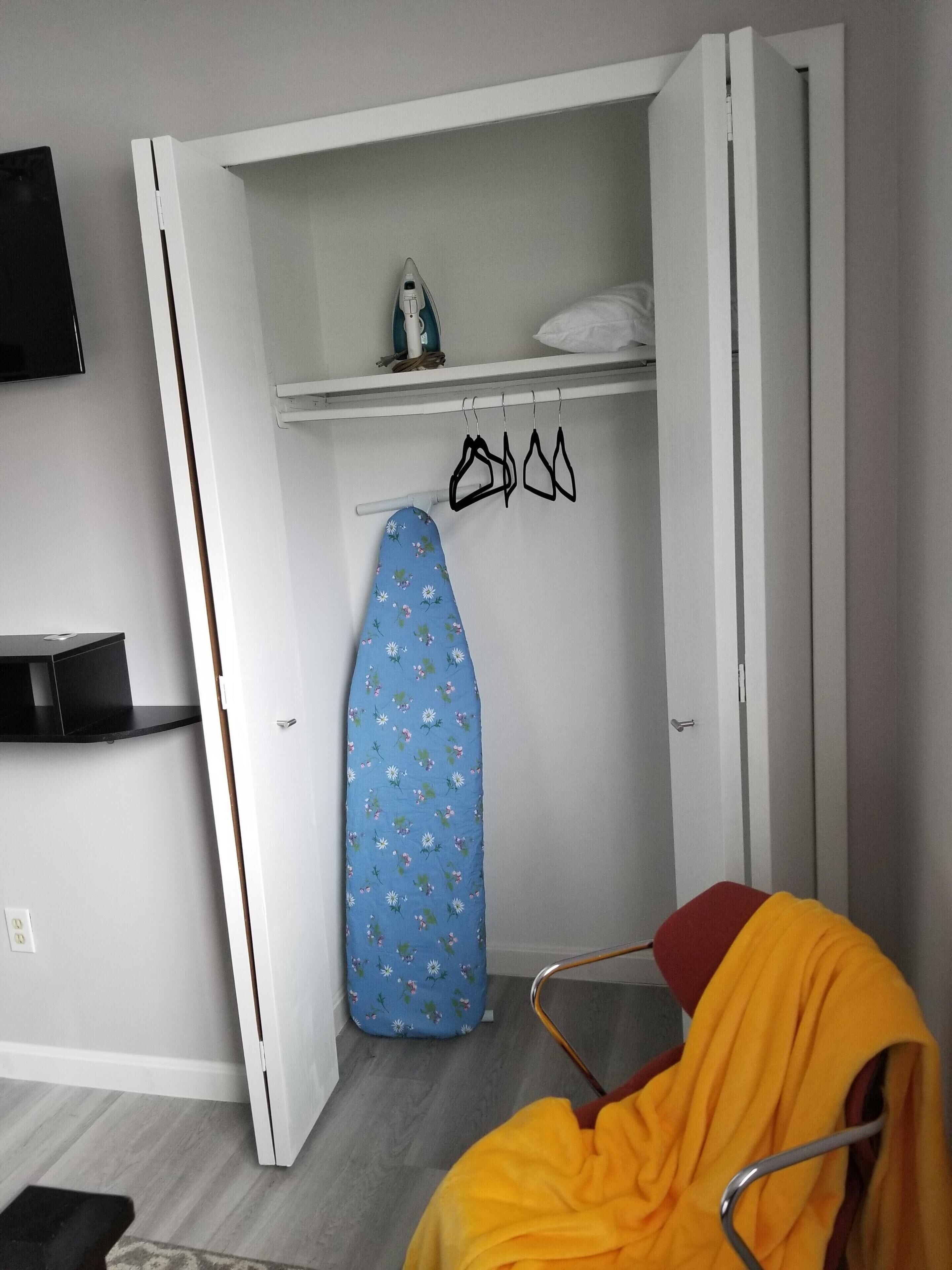A compact room with a bed, a chair, a wardrobe, and a hanging iron board.