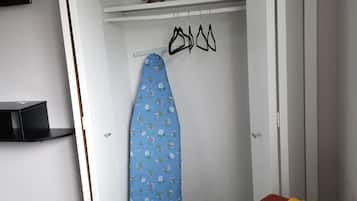 A compact room with a bed, a chair, a wardrobe, and a hanging iron board.