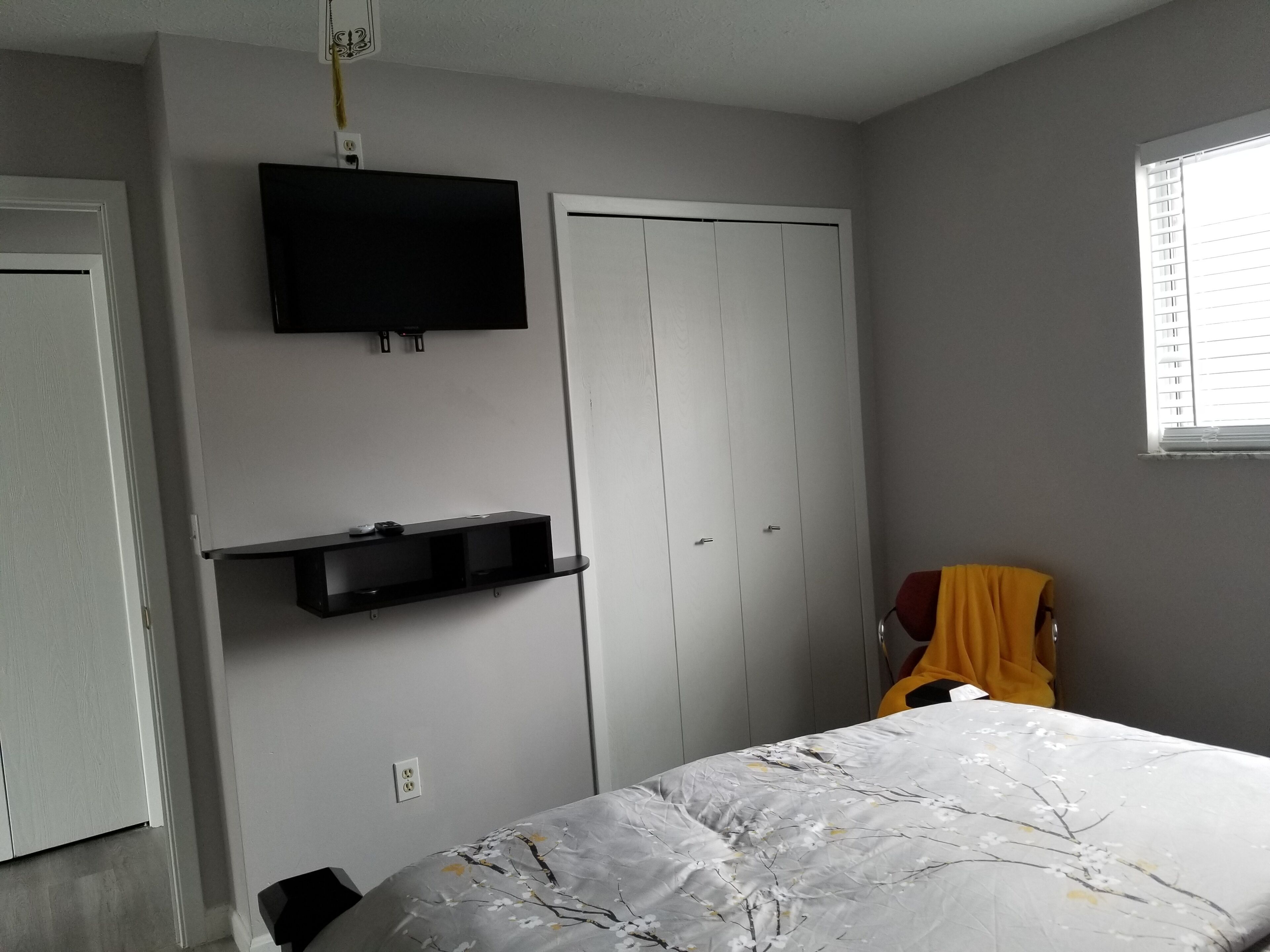 Un dormitorio con cama, televisión montada en la pared, armario y ventana con persianas.