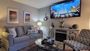 Ein Wohnzimmer mit grauem Sofa, zwei gerahmten Kunstwerken, einem Fernseher der eine Stadt zeigt, einer Uhr und einem gemusterten Sessel.