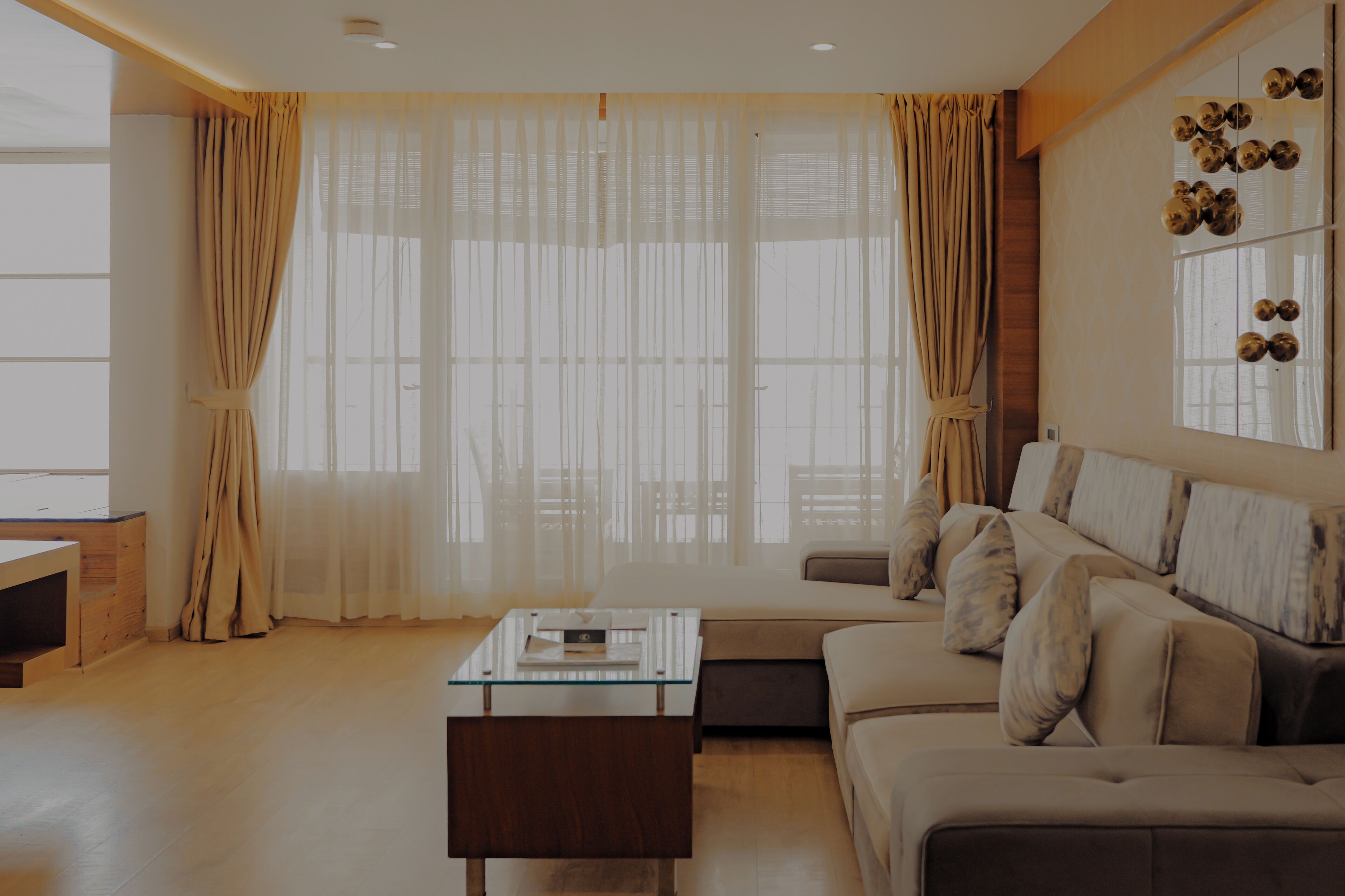 A modern living room with a glass coffee table, a sofa, and a large window with sheer curtains.