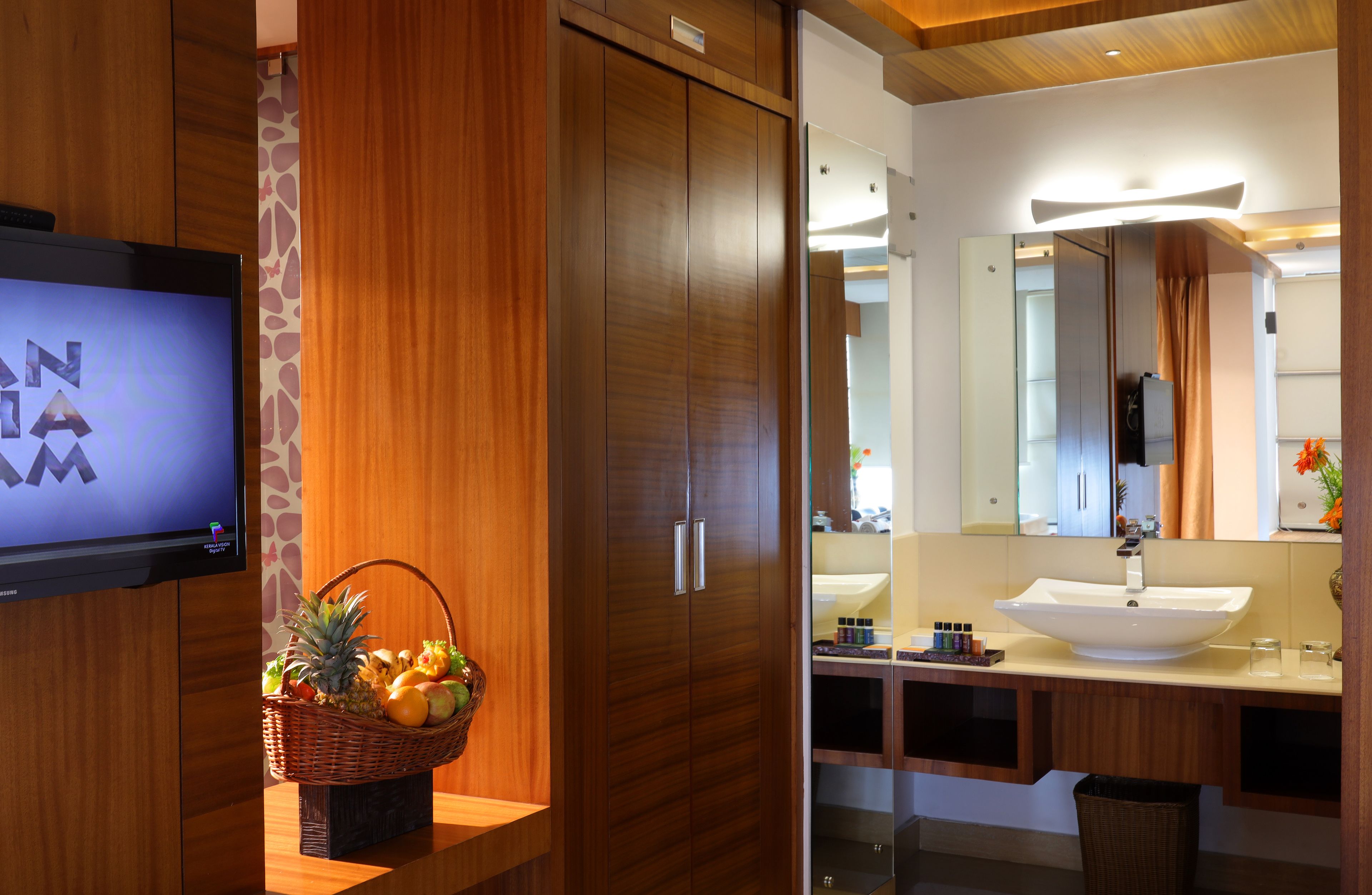 Une salle de bain d’hôtel avec un meuble-lavabo en bois, un grand miroir, un lavabo et un téléviseur diffusant l’émission « NAMA ».