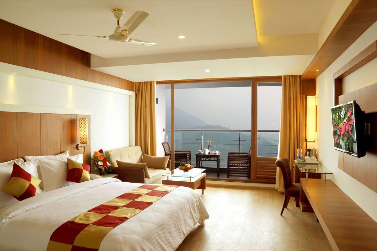 A hotel room with a large bed, a desk with a chair, a television, and a balcony with a view of the sea.