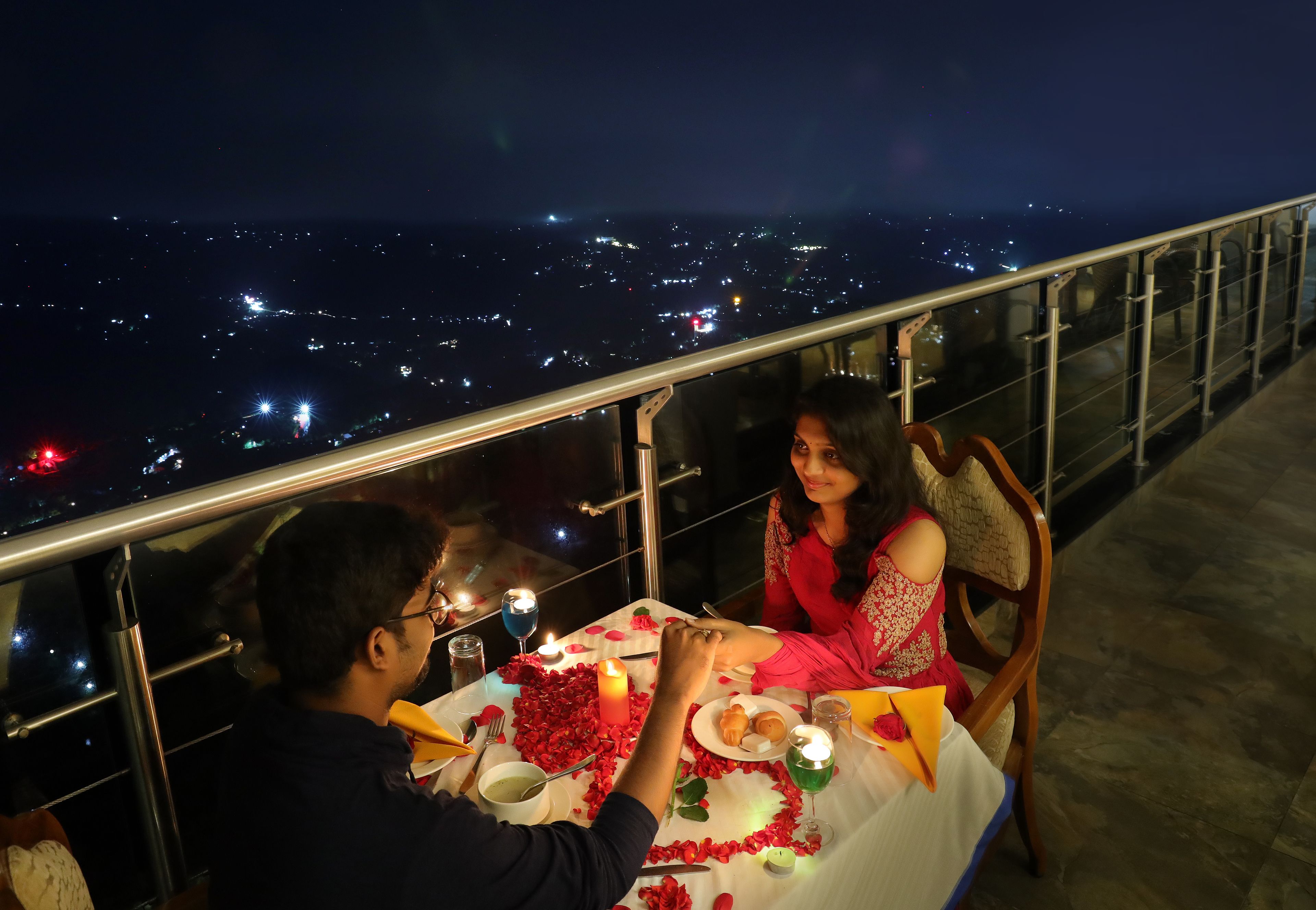 A couple dining at a table with a city view, surrounded by rose petals and candles.