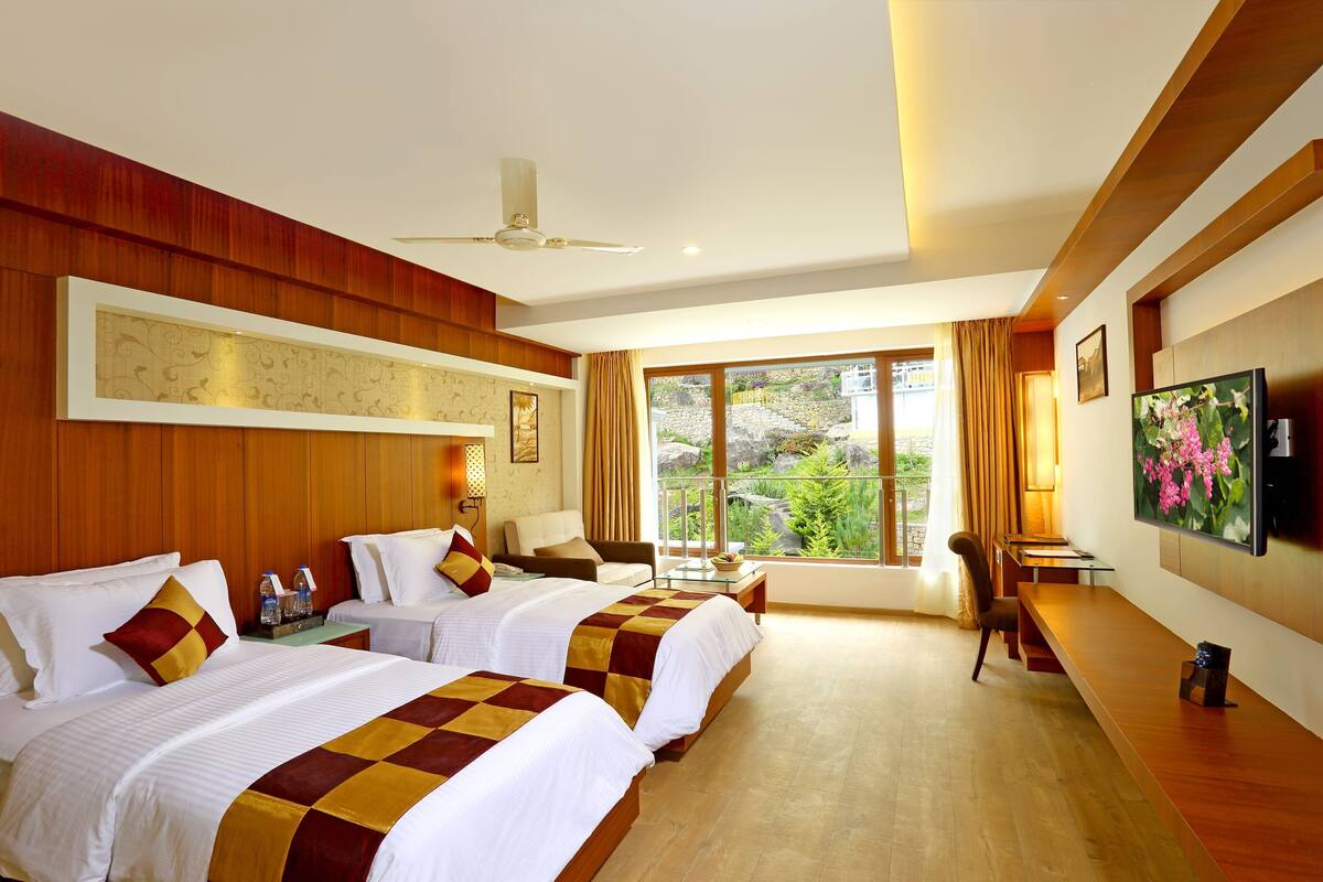 A hotel room with two beds, a ceiling fan, a TV, and a large window with a view of greenery.