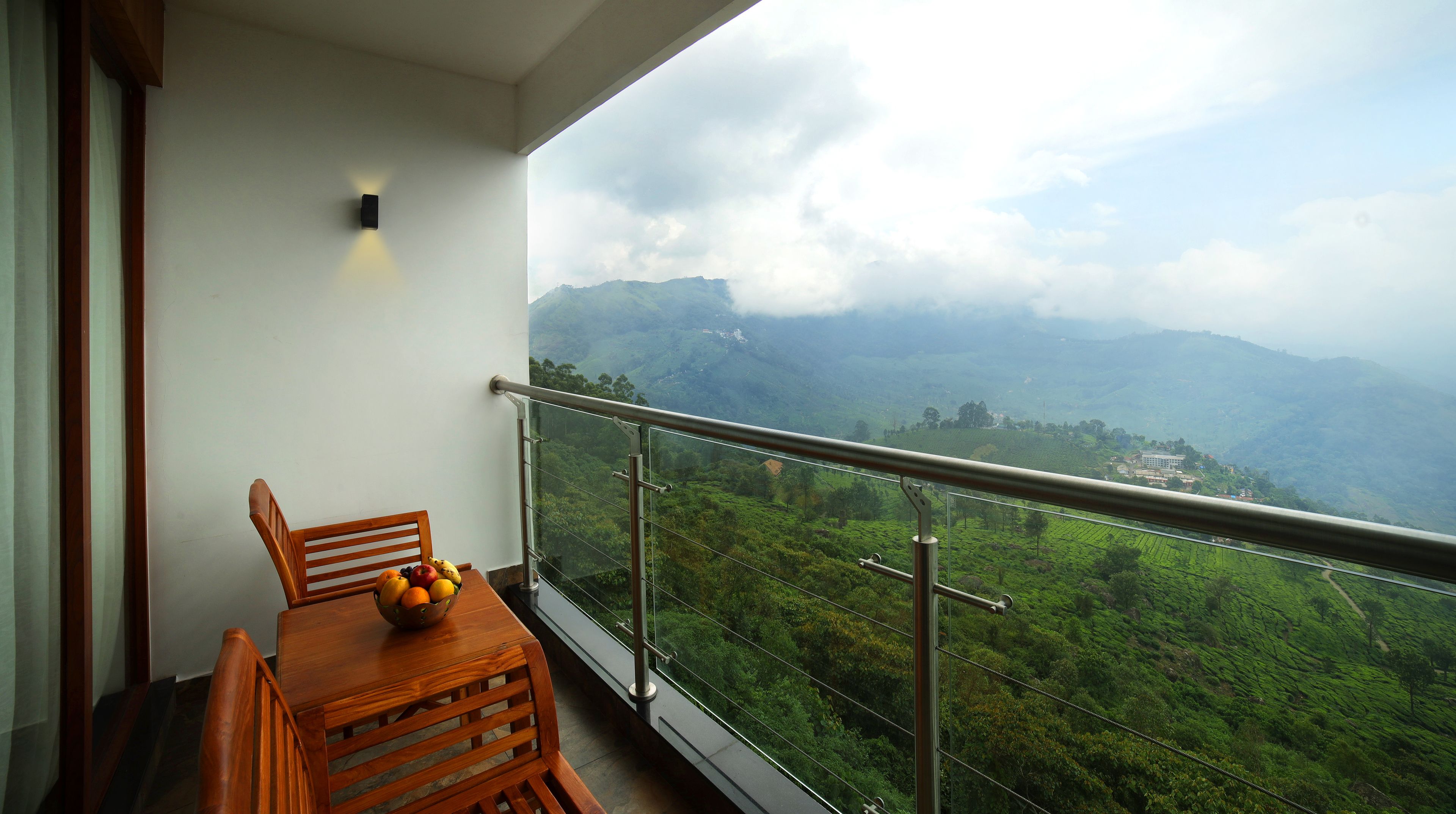 A balcony with a wooden table and chairs, a bowl of fruit, and a view of lush green hills and mountains.