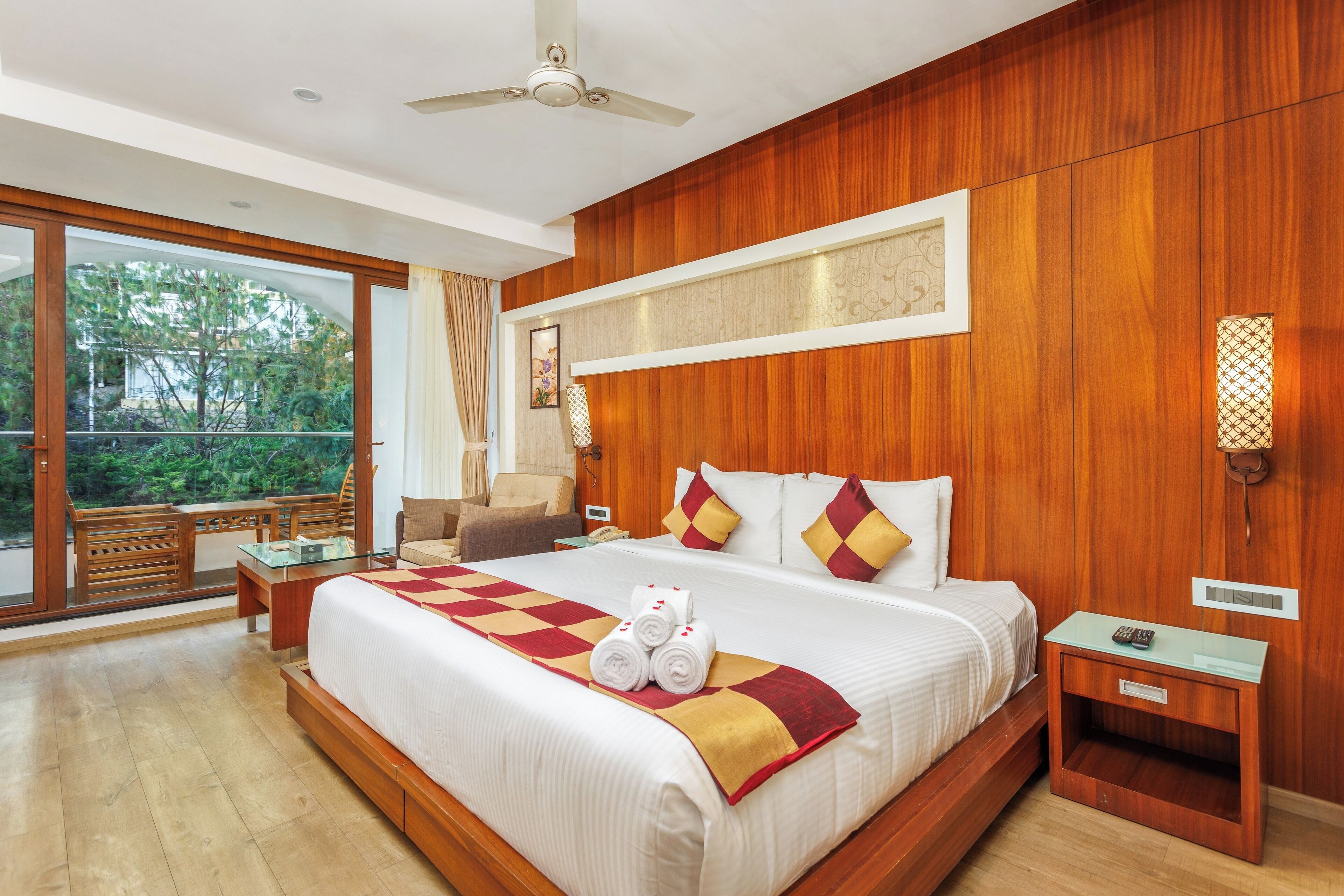 A hotel room with a large bed, wooden paneling, a ceiling fan, a nightstand, and a view through a glass door.