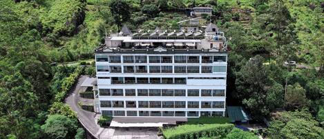 A multi-story hotel with a flat roof, surrounded by lush greenery and hills.