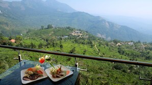 Una mesa con comida y bebidas, con vista a un paisaje montañoso con plantaciones de té y un edificio distante.