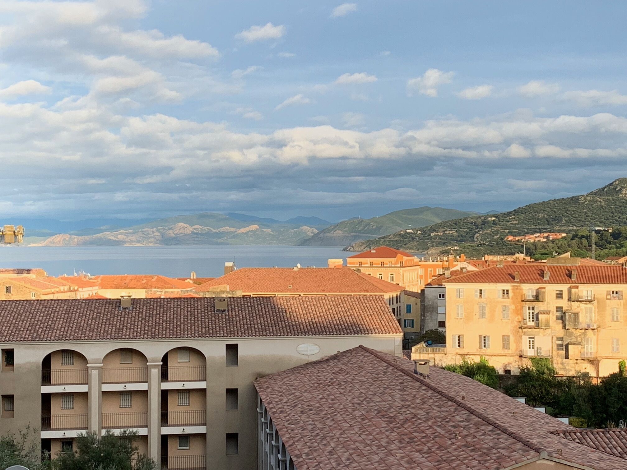 A coastal town with terracotta-roofed buildings, a sea view, and a mountainous backdrop.