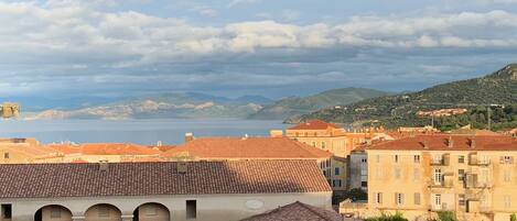 A coastal town with terracotta-roofed buildings, a sea view, and a mountainous backdrop.