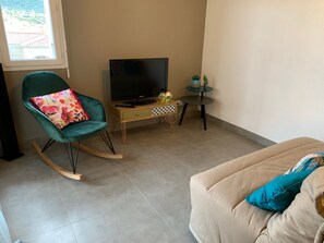 A living room with a teal rocking chair, a beige sofa, a television on a wooden stand, and a small round table with a glass top. - Apartment/ flat - Ile Rousse (Ile Rousse)