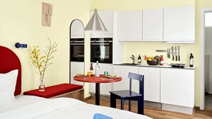 A compact hotel room with a bed, a small dining table with a chair, a kitchenette, and a red bench.