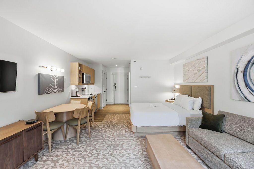 Ett hotellrum med en säng, en soffa, ett skrivbord, en stol, en TV och ett litet kök.