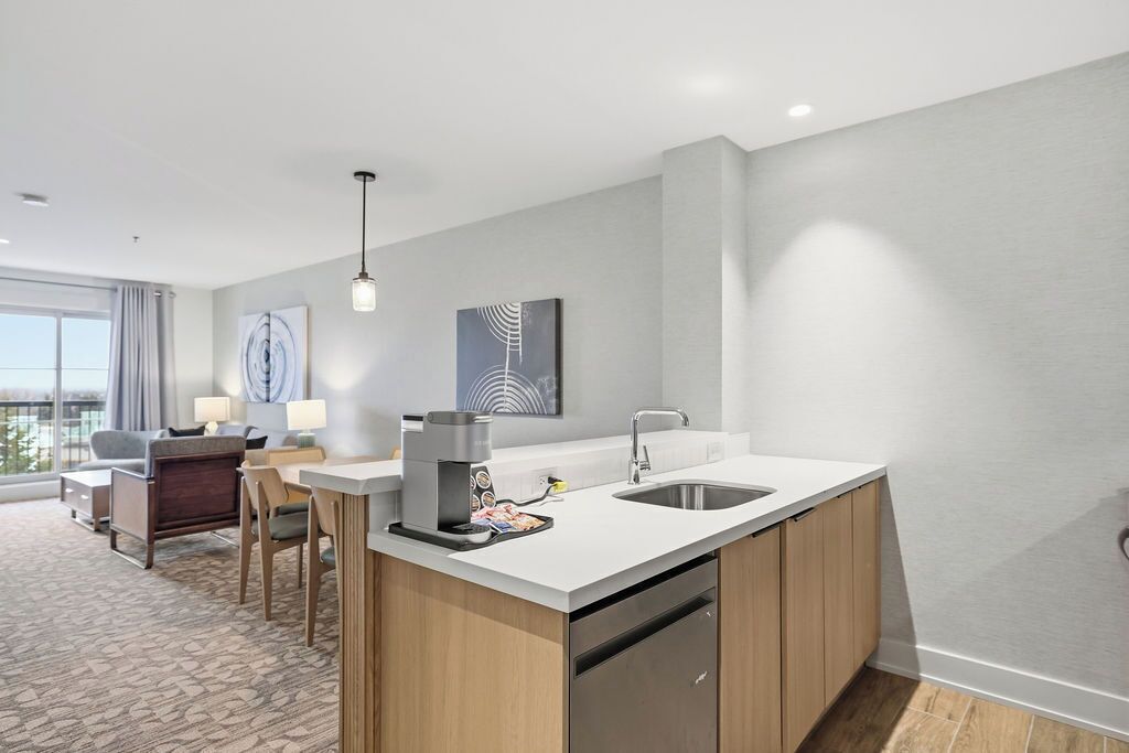 A modern hotel room with a kitchenette, a dining area with a sofa and chairs, and artwork on the walls.