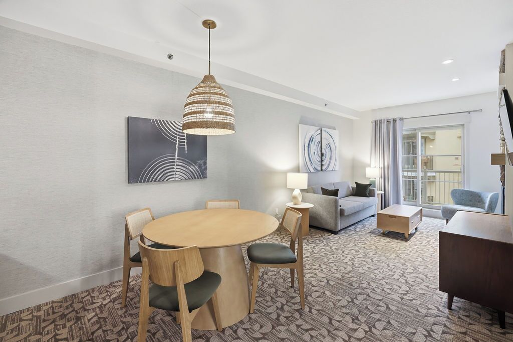 A hotel room with a round dining table, chairs, a sofa, a lamp, and a large window with curtains.