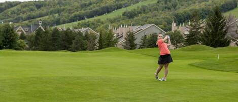 Un terrain de golf avec un joueur portant un maillot rouge et une jupe noire, des maisons en arrière-plan et un paysage vallonné.