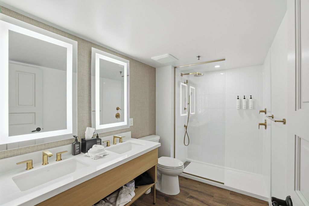 A modern bathroom with a large mirror, two sinks, a toilet, and a walk-in shower.