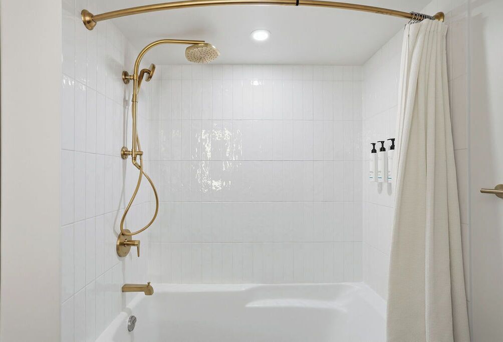 A shower with a gold showerhead and curved rail, a white bathtub, and a white curtain.