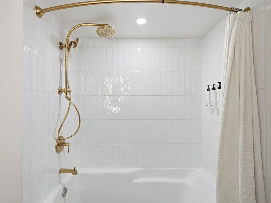 Ducha con cabezal dorado y riel curvo, bañera blanca y cortina blanca.