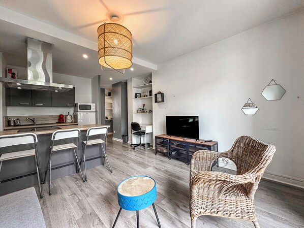 A modern living room with a TV, wicker chairs, a blue stool, and a kitchen area with a microwave.