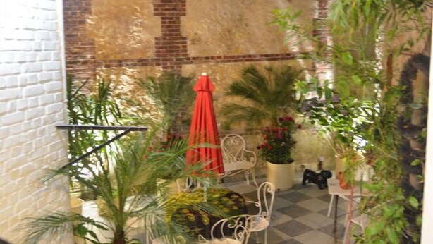 Une cour dallée de motifs à carreaux, agrémentée de plantes en pot, d’un parasol rouge et de chaises en métal blanc.