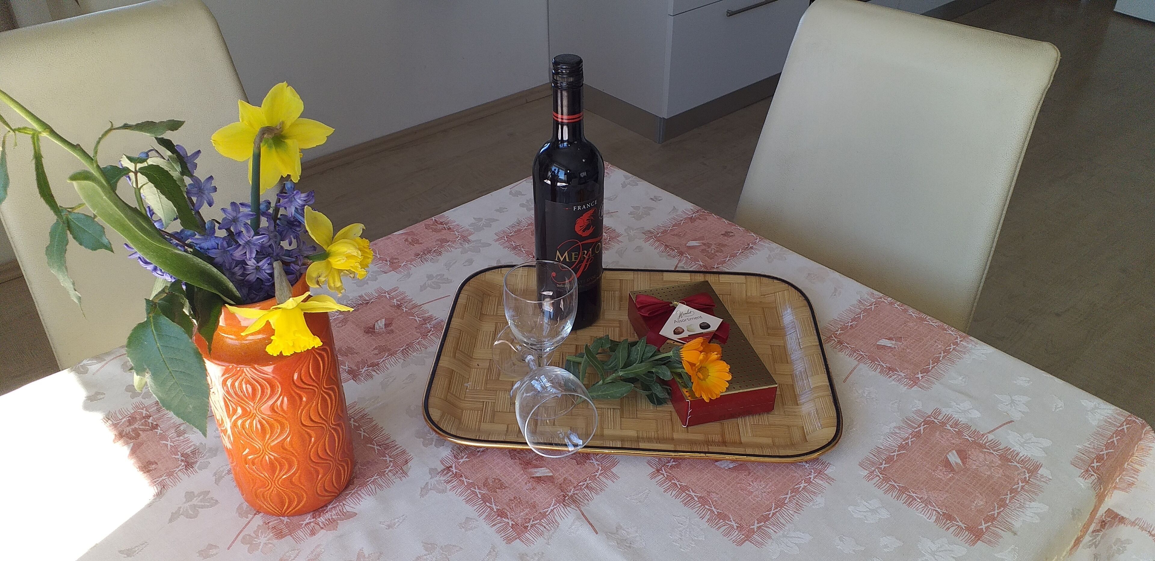 Uma mesa de jantar com um vaso de flores, uma garrafa de vinho e uma pequena caixa de presente.