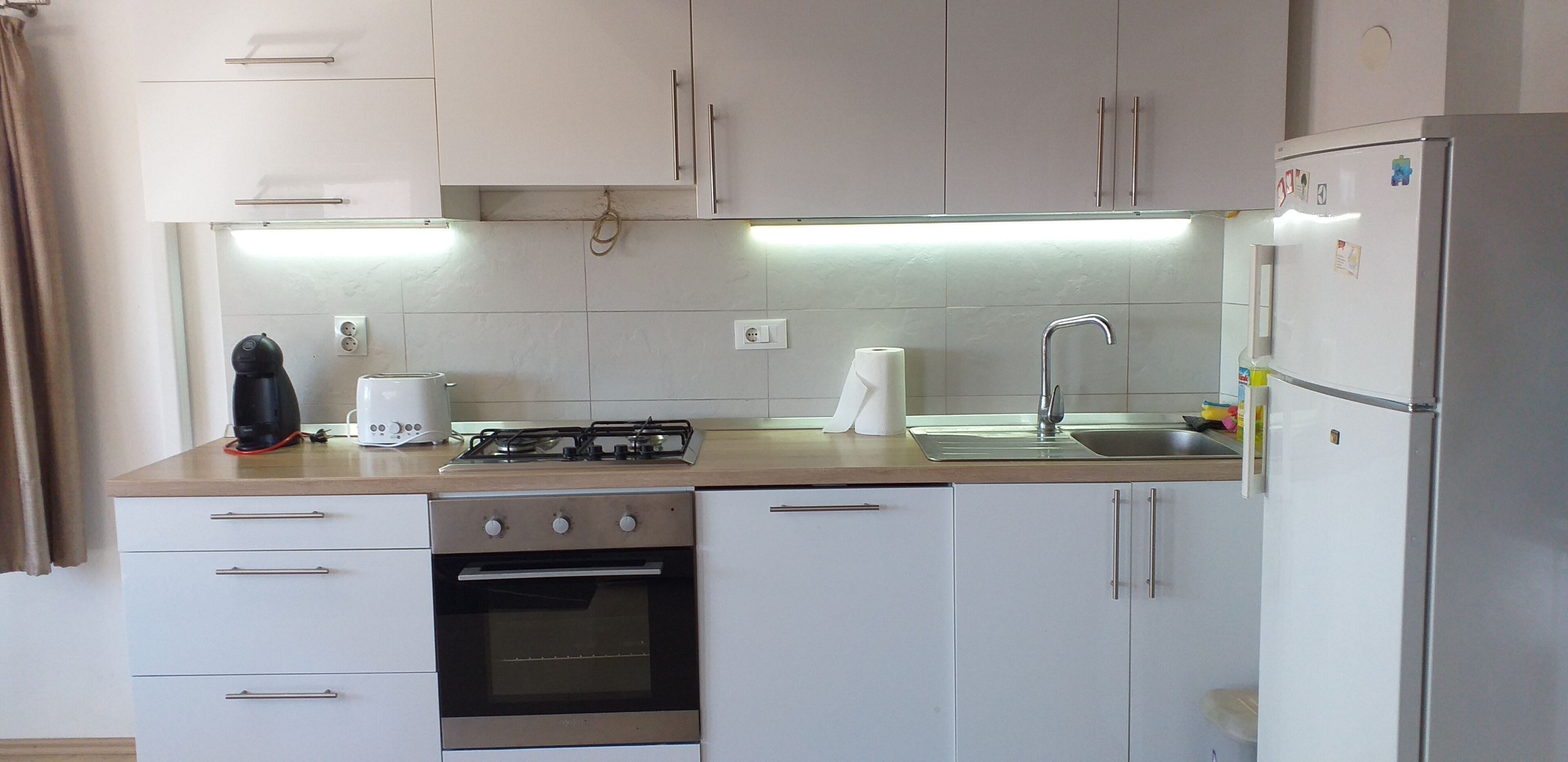 Une cuisine de taille réduite, avec des meubles blancs, une cuisinière à gaz, un évier et un réfrigérateur.