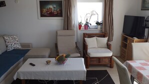 Een woonkamer met een bank, een houten stoel, een kleine tafel met fruit, een televisie en potplanten bij een raam.