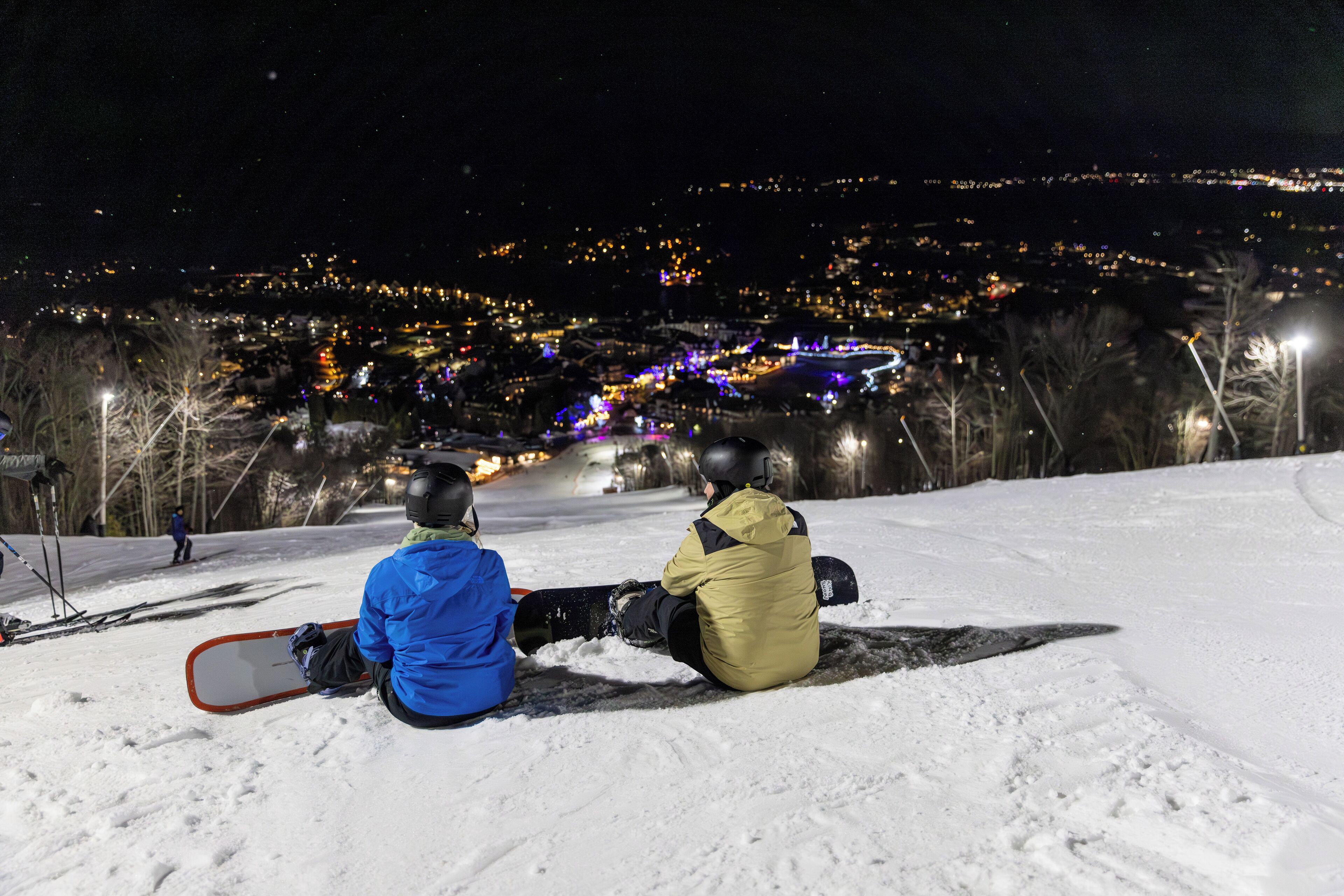 Zwei Personen sitzen auf einem verschneiten Hang mit Blick auf eine Stadt bei Nacht.