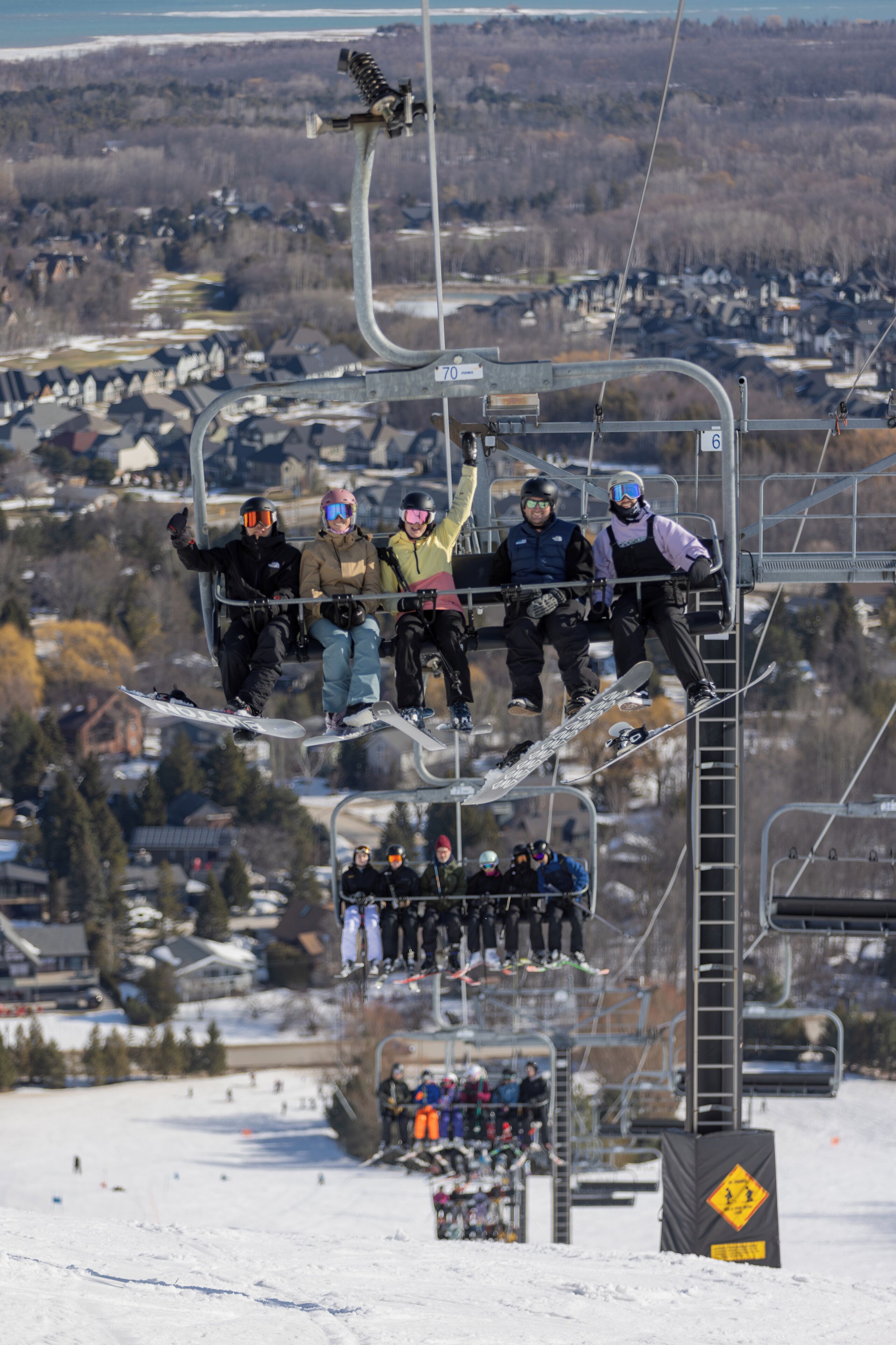 A ski lift with passengers seated on it, overlooking a snowy landscape with buildings in the background.