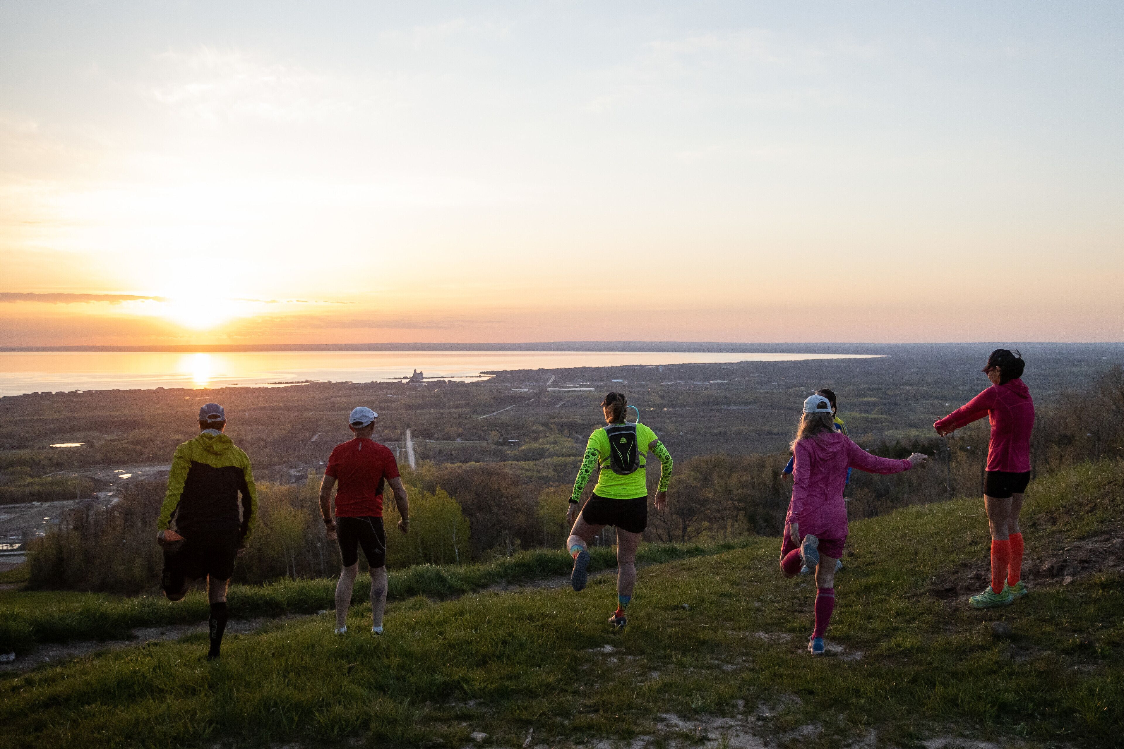 Eine Gruppe von Menschen läuft einen grasbewachsenen Hügel hinauf, im Hintergrund ein Sonnenuntergang.