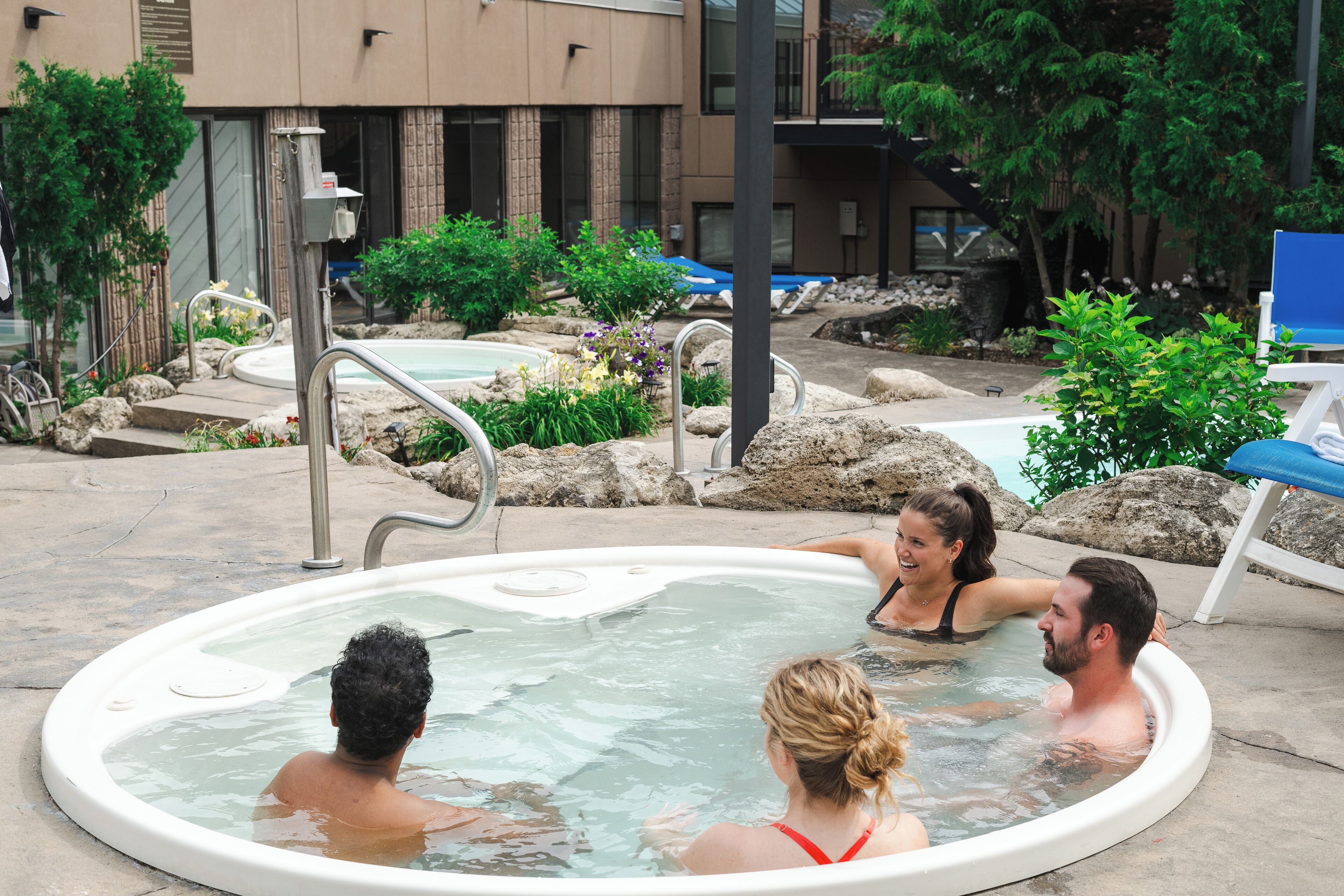 A hot tub area with a pool, surrounded by rocks and plants, and a building in the background.