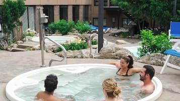 Un área de hidromasaje con piscina, rodeada de rocas y plantas, y un edificio al fondo.