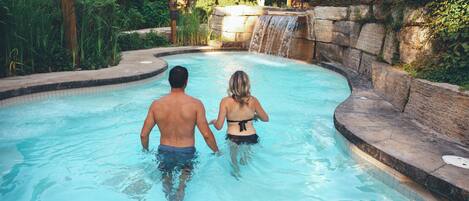 A man and a woman walking in a pool with a waterfall feature.