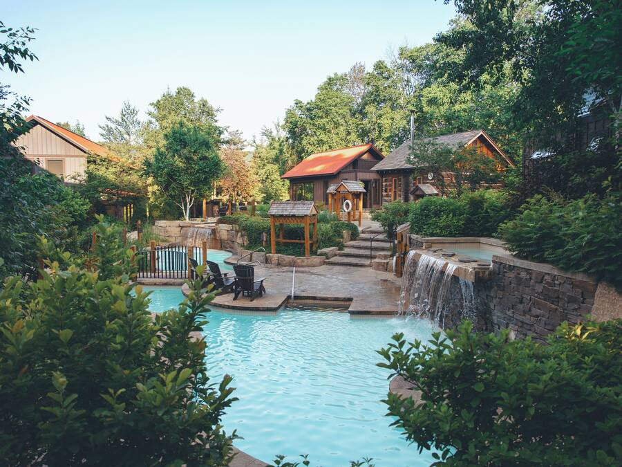 A pool area with a waterfall, surrounded by lush greenery and wooden cabins.
