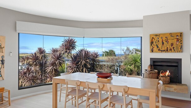 A modern dining area with a large window overlooking a garden, a fireplace, and a painting on the wall. - Beautiful spacious pet friendly home next to the ocean (Smiths Beach)